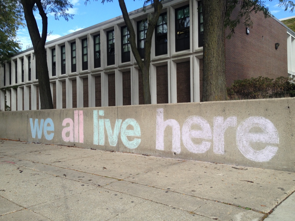 The words "we all live here" written in chalk on a wall in front of a building